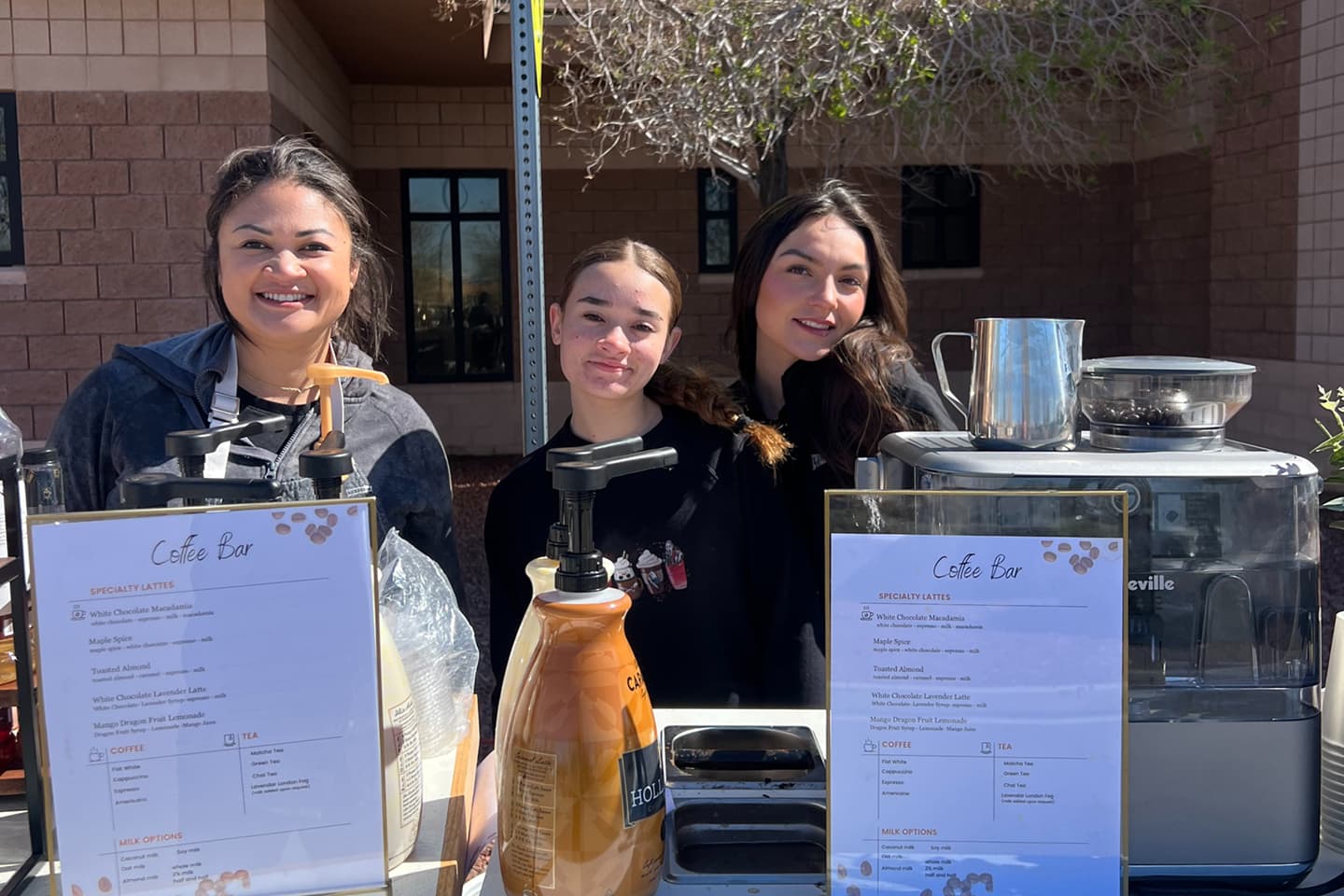 Three All That & A Latte baristas pose behind a coffee bar and gourmet beverage menu