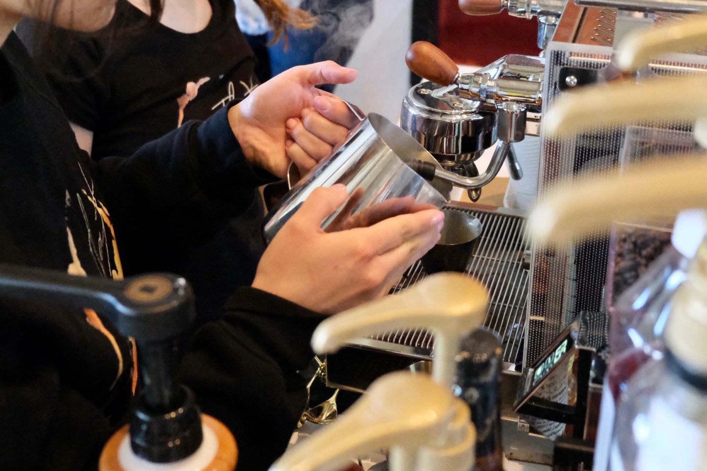 Metal cup in hands being used with espresso machine near syrup pumps.